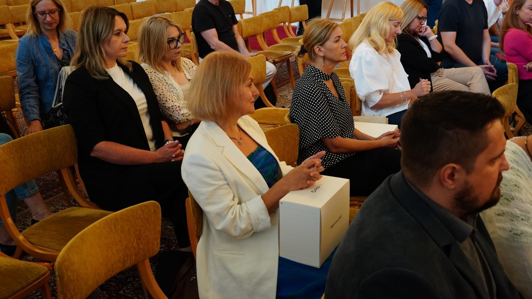 Sala Balowa, siedzący słuchacze - kobiety i mężczyźni. Jedną z osób jest kierownik DEM Edyta Kucaba-Łyszczek, która trzyma na kolanach pudło z goglami VR