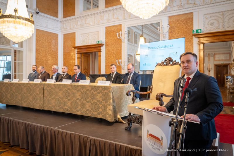 Sala Balowa, dyrektor dr Piotr Szopa mówi do zebranych, stoi. Za nim stół i uczestnicy podpisujący porozumienie Doliny Sarmackiej (7 mężczyzn)