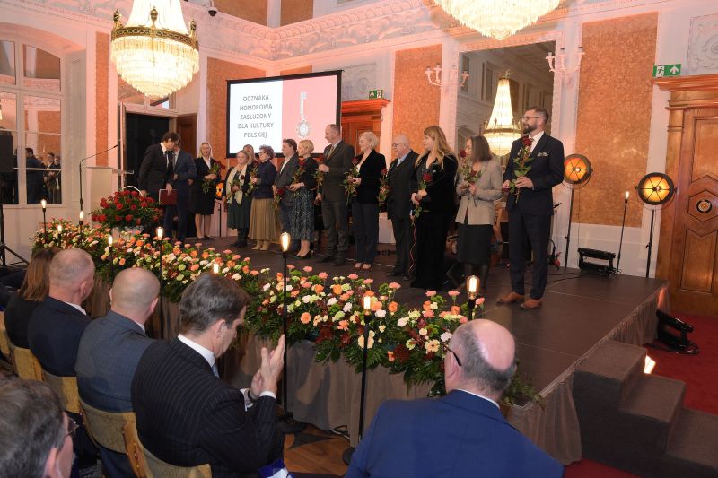 Odznaczeni odznakami honorowymi na scenie - 10 nagrodzonych, prowadzący, dyr. P. Szpanowski, asystentki do wręczania kwiatów. Odznaczeni trzymają medale i czerwone róże