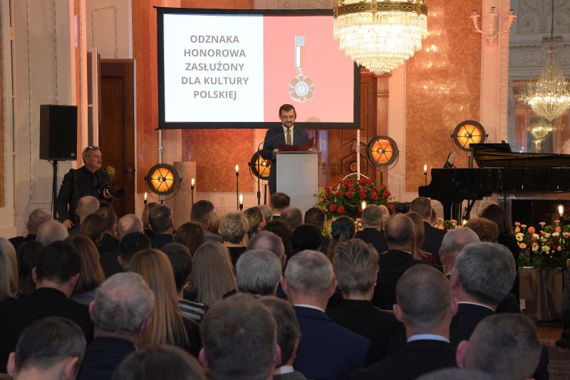 Sala Balowa, goście wydarzenia siedzą na krzesłach, przy mównicy dyrektor Piotr Szpanowski