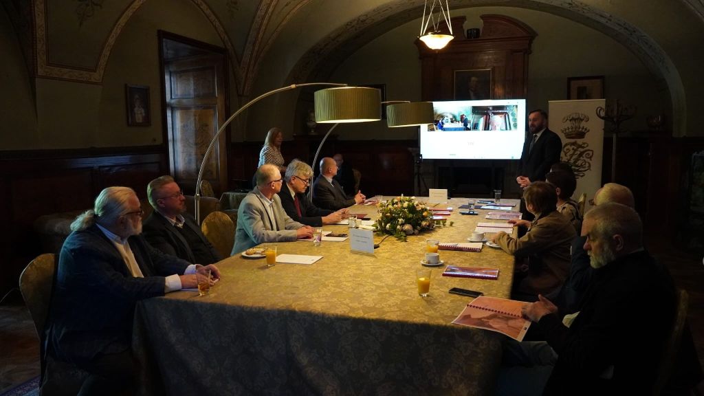 Zdjęcie przedstawia inne ujęcie posiedzenia Rady Muzeum w Zamku w Łańcucie. Grupa osób siedzi wokół dużego stołu nakrytego ozdobnym obrusem, na którym znajdują się materiały konferencyjne, napoje oraz kompozycja kwiatowa. W tle widoczny jest ekran z uczestnikami połączenia online oraz stojący obok mężczyzna prowadzący spotkanie. Sala o kolebkowym sklepieniu z dekoracyjnymi malowidłami jest oświetlona przez stylowe lampy o nowoczesnym kształcie. Panuje atmosfera skupienia i profesjonalizmu.