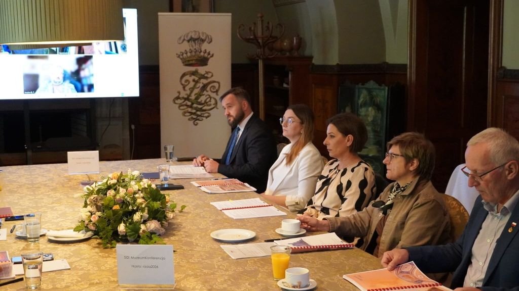 Zdjęcie przedstawia zbliżenie na grupę pięciu osób siedzących po jednej stronie stołu podczas posiedzenia Rady Muzeum w Zamku w Łańcucie. Uczestnicy, ubrani w stroje biznesowe, uważnie słuchają lub przeglądają dokumenty. Na stole przed nimi znajdują się filiżanki, szklanka z sokiem oraz kolorowy bukiet róż. W tle, obok ekranu z podglądem wideokonferencji, stoi biały baner z herbem zamku. Na pierwszym planie widoczna jest plakietka z danymi do logowania do sieci Wi-Fi. Panuje atmosfera merytorycznej dyskusji w zabytkowym wnętrzu.