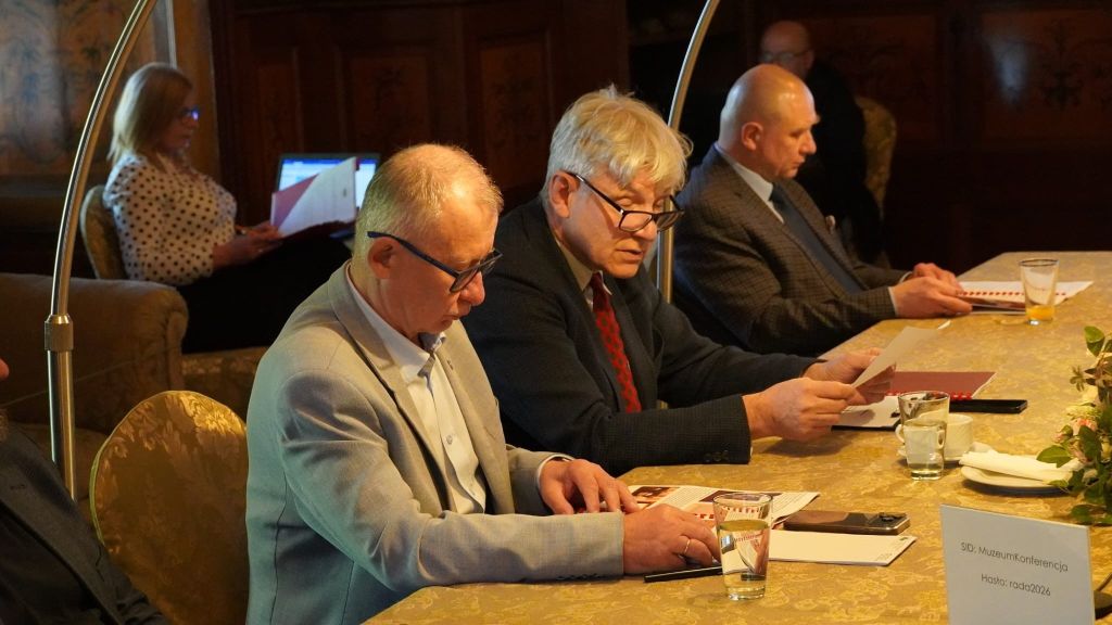 Zdjęcie przedstawia zbliżenie na trzech mężczyzn siedzących przy stole konferencyjnym podczas posiedzenia Rady Muzeum. Uczestnicy są skupieni na przeglądaniu dokumentów i materiałów drukowanych. W tle, po lewej stronie, widać kobietę pracującą przy laptopie. Na stole, przed uczestnikami, znajdują się szklanki z napojami oraz plakietka z danymi dostępowymi do sieci Wi-Fi. Scena ma miejsce w historycznym, nastrojowym wnętrzu Zamku w Łańcucie, co podkreślają ozdobne obicia krzeseł i detale w tle.