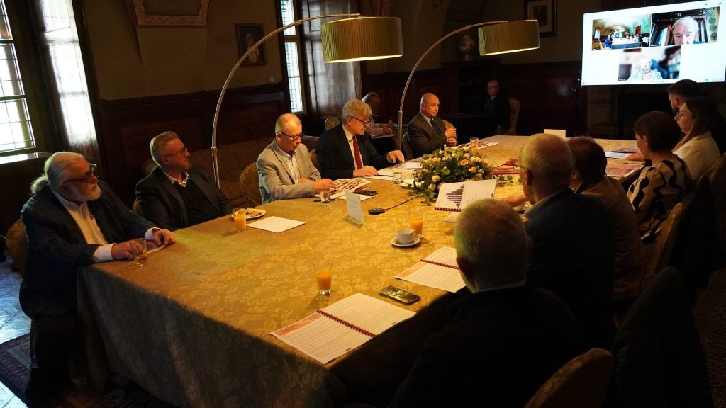 Zdjęcie przedstawia szeroki kadr posiedzenia Rady Muzeum w Zamku w Łańcucie. Uczestnicy siedzą przy dużym, oświetlonym stole, analizując rozłożone dokumenty i wykresy. W głębi sali widoczny jest ekran multimedialny z uczestnikami biorącymi udział w spotkaniu online. Historyczny charakter wnętrza podkreślają sklepienia z dekoracjami, stylowe meble oraz wpadające przez wysokie okna naturalne światło, które miesza się z ciepłym blaskiem nowoczesnych lamp łukowych.