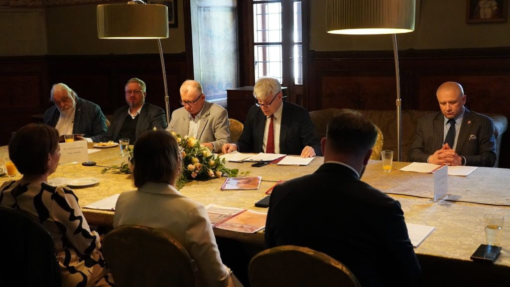 Zdjęcie przedstawia grupę osób siedzących wokół dużego stołu podczas posiedzenia Rady Muzeum w Zamku w Łańcucie. Centralną część kadru zajmuje pięciu mężczyzn w strojach wizytowych, skupionych na dokumentach i dyskusji. Na pierwszym planie widoczne są sylwetki trzech kolejnych osób siedzących tyłem do obiektywu. Stół zdobi elegancka kompozycja z jasnych róż, a całe spotkanie odbywa się w nastrojowo oświetlonym, zabytkowym wnętrzu z wysokimi oknami i dekoracyjnymi ścianami. Dwie nowoczesne lampy podłogowe doświetlają blat stołu, podkreślając profesjonalny charakter obrad.