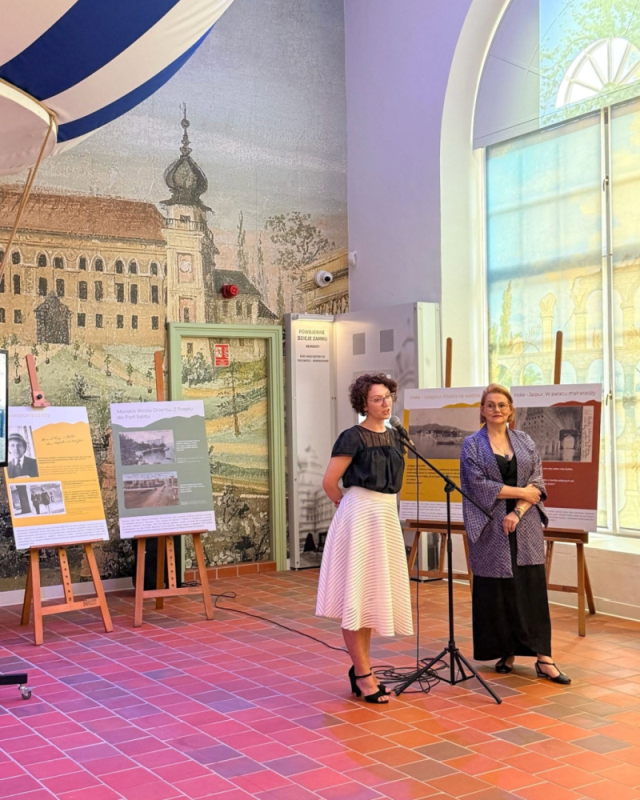 Scena ogólna: Zdjęcie przedstawia wernisaż wystawy w Muzeum – Zamek w Łańcucie. W jasnym pomieszczeniu, na tle fototapety z motywem architektonicznym, stoją dwie kobiety.  Postacie:  Po lewej: Kobieta w okularach, o ciemnych, kręconych włosach, ubrana w czarną bluzkę i białą, plisowaną spódnicę, przemawia do mikrofonu na statywie.  Po prawej: Blondynka w okularach, ubrana w czarne spodnie i wzorzystą, fioletowo-niebieską narzutkę, słucha wypowiedzi z rękami splecionymi przed sobą.  Otoczenie: Po lewej stronie widoczny jest fragment pasiastego, biało-niebieskiego zadaszenia. Za kobietami, na drewnianych sztalugach, stoją tablice informacyjne z tekstem i grafikami (m.in. z napisami „Morskie Wrota Orientu…” i „Indie – Jaipur…”). Po prawej stronie znajduje się duże, półokrągłe okno, przez które wpada jasne światło, a na podłodze ułożona jest czerwona terakota.