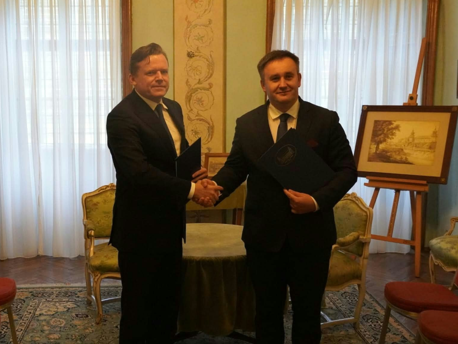 Na zdjęciu widocznych jest dwóch elegancko ubranych mężczyzn w garniturach, stojących w klasycznie urządzonej sali i ściskających sobie dłonie. Mężczyzna po lewej stronie trzyma w drugiej ręce ciemną teczkę, a mężczyzna po prawej przytrzymuje niebieską teczkę z dokumentami. Obaj patrzą w stronę aparatu z uśmiechem. W tle znajduje się okrągły stolik nakryty zielonym obrusem oraz stylizowane krzesła z jasną tapicerką. W pokoju stoją również inne krzesła z czerwonym obiciem, a pod ścianą na sztalugie wyeksponowany jest oprawiony w ramę obraz. Okna w tle przesłonięte są jasnymi, zwiewnymi zasłonami, przez które wpada naturalne światło. Całość ma oficjalny, uroczysty charakter.