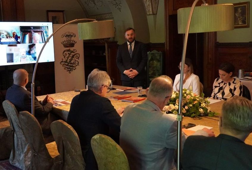 Zdjęcie przedstawia posiedzenie Rady Muzeum w zabytkowym wnętrzu Zamku w Łańcucie. Grupa osób siedzi przy długim stole ozdobionym kwiatami. U szczytu stołu stoi przemawiający mężczyzna w garniturze. Po lewej stronie umieszczono duży monitor z widocznym podglądem wideokonferencji oraz baner z herbem zamku. Wnętrze charakteryzuje się historycznym wystrojem, sklepieniami z polichromiami oraz stylowym, ciemnym umeblowaniem. Spotkanie łączy formę stacjonarną z uczestnictwem zdalnym.