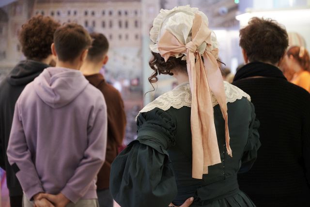 Dawna Oranżeria, młodzież odwrócona tyłem do aparatu, ogląda ekspozycję. Za nimi tyłem do aparatu kobieta w stroju z epoki (w nakryciu głowy, w zielonej sukni z kołnierzem)